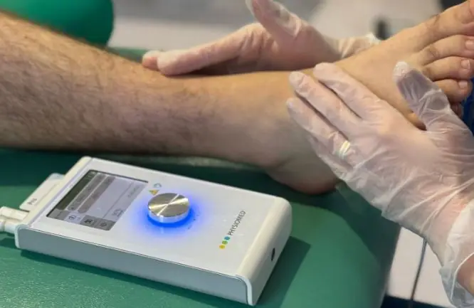Terapie Deep Oscillation: aparatul Physiomed pe masă și mâinile terapeutului cu mănuși aplicate pe gleznă – Clinica Kinetoterapia Piatra-Neamț